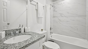Un baño con lavamanos de encimera de granito, inodoro blanco y bañera con cortina de ducha.