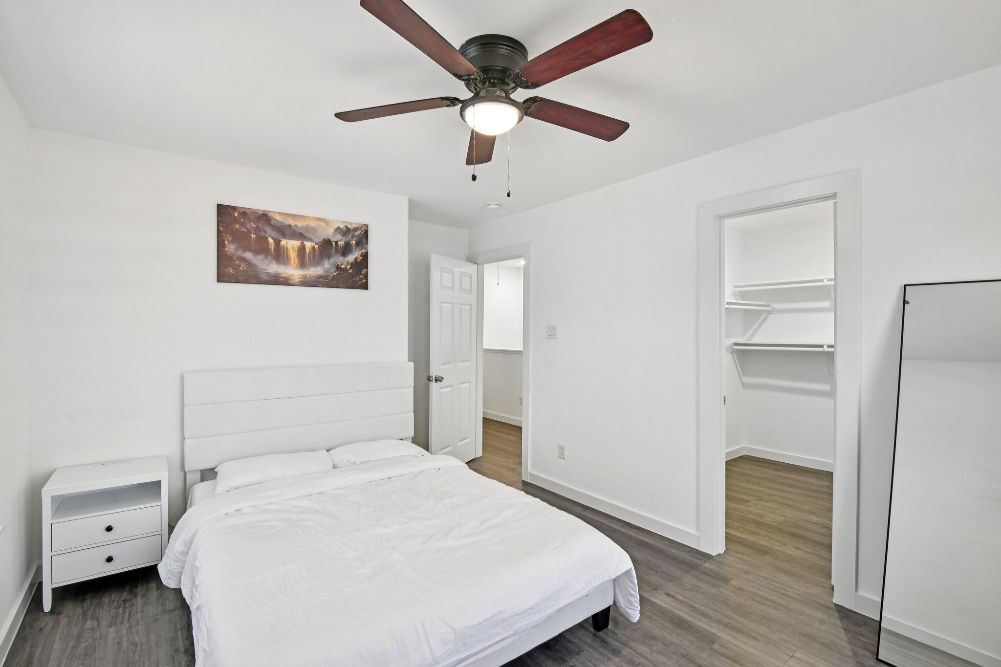 Ein Schlafzimmer mit Bett, Nachttisch, Deckenventilator, einem Gemälde und einem begehbaren Kleiderschrank.