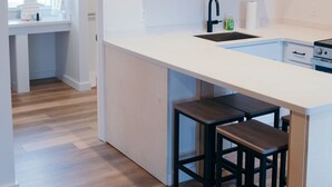 Eine moderne Küche mit weißer Arbeitsplatte, schwarzen Barhocker, Holzboden und einem Fenster mit Handtuchhalter.
