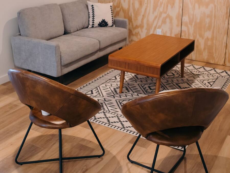Una sala de estar moderna con un sofá gris, una mesa de centro de madera, dos sillones marrones y una alfombra estampada.
