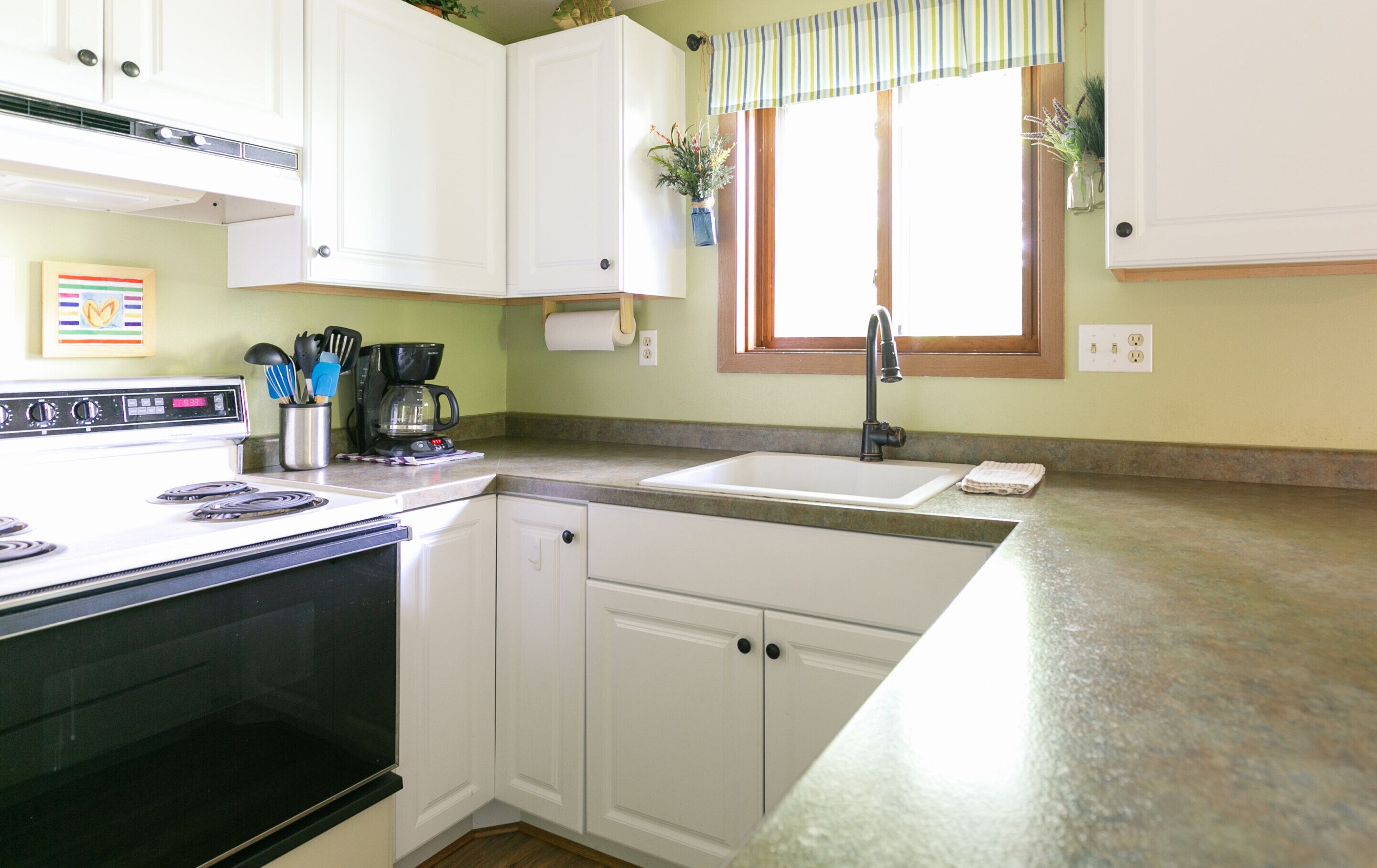 A kitchen with white cabinets, a stove, a coffee maker, and a window with a plant.