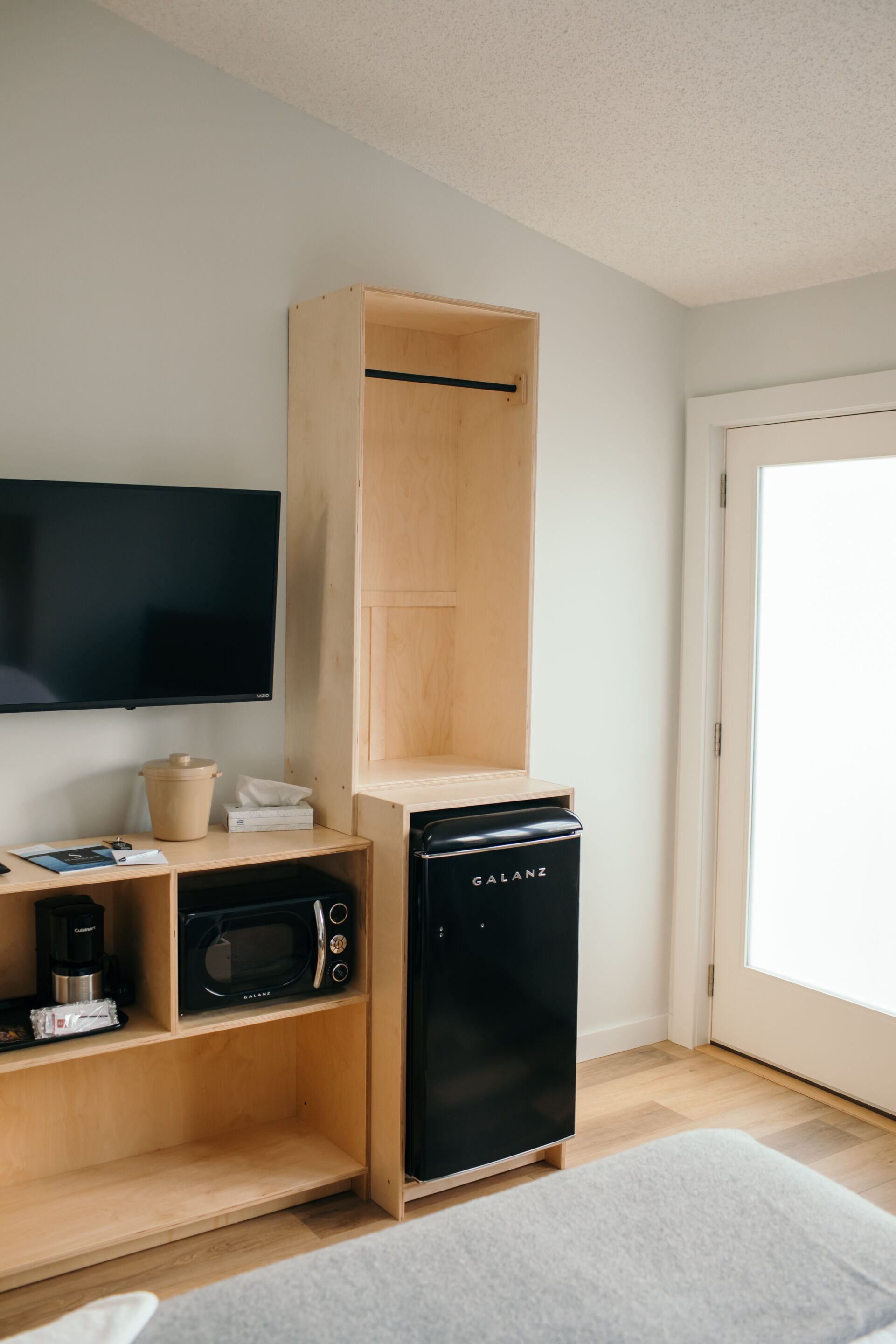 Una kitchenette compacta con microondas, cafetera y un refrigerador negro. Un mueble de madera está integrado en la pared, y un televisor de pantalla plana está montado encima.
