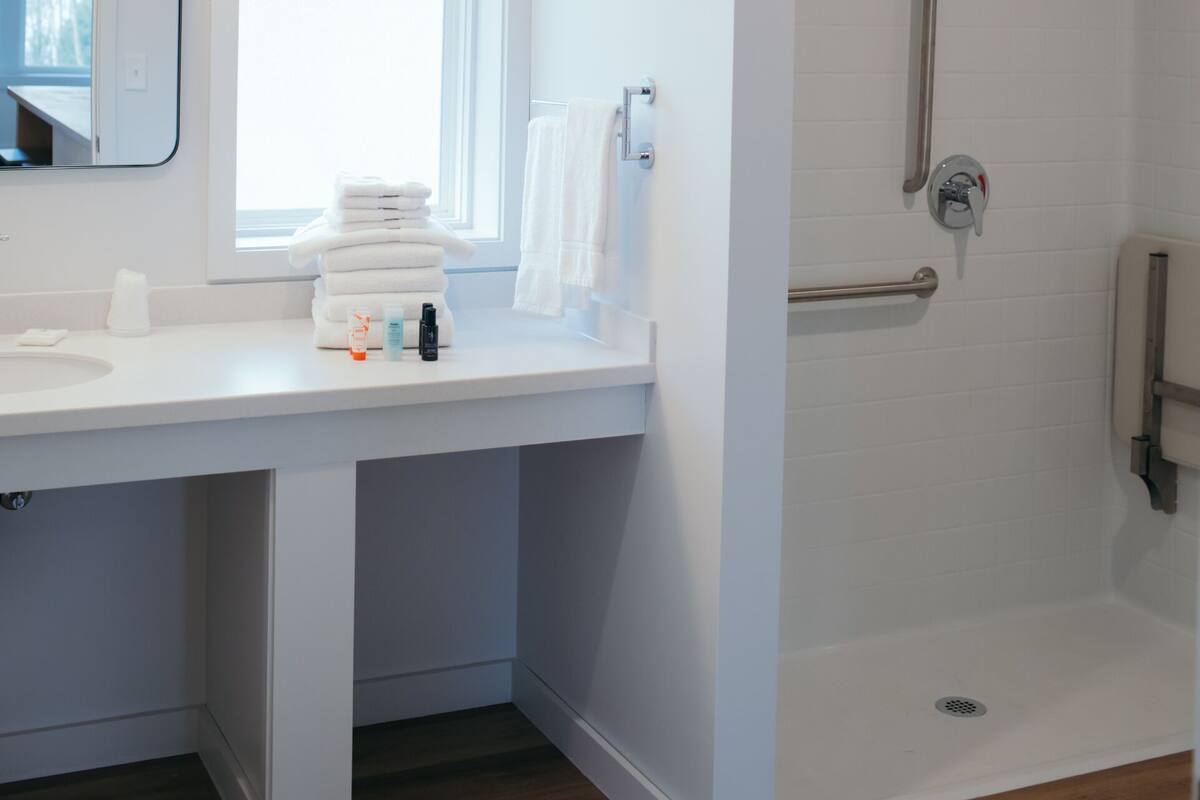 A modern bathroom with a white vanity, a mirror, a window, and a walk-in shower with a handrail.