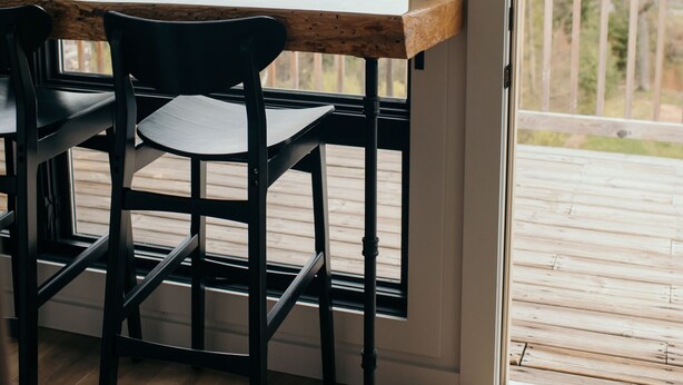 Ein Zimmer mit Holzterrasse, die durch eine Glastür sichtbar ist, einem Holztisch und zwei schwarzen Stühlen.