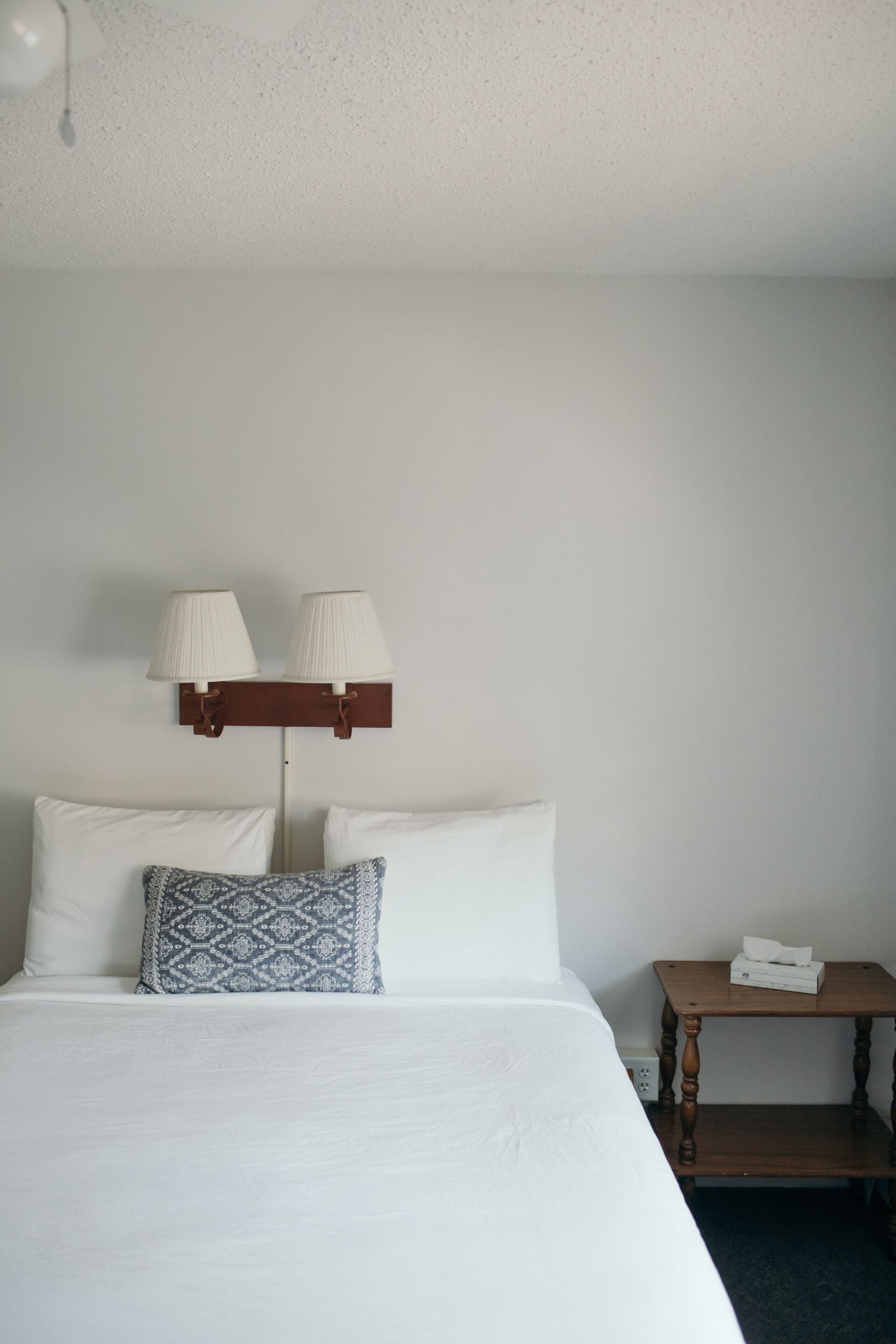 Una cama bien hecha con sábanas blancas, un cojín estampado y dos lámparas fijadas a la pared.