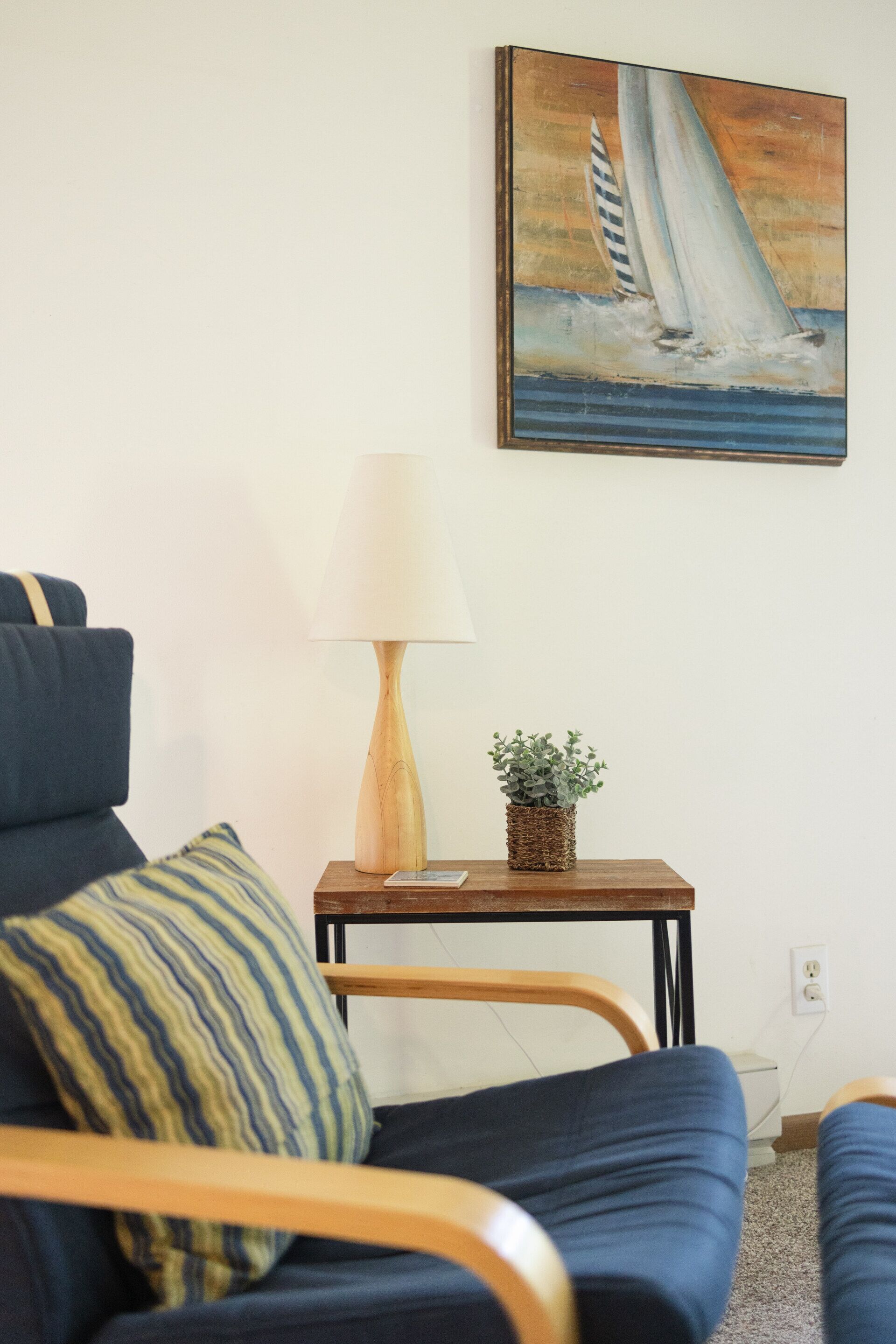 A room with a blue sofa, a wooden side table with a lamp and a small plant, and a painting of a sailboat on the wall.