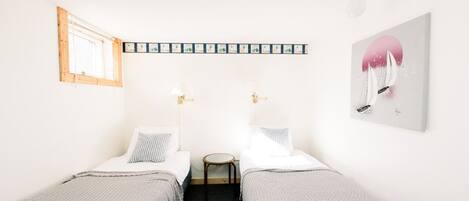 A small, clean room with two single beds, a small table, a lamp, and a framed picture on the wall.