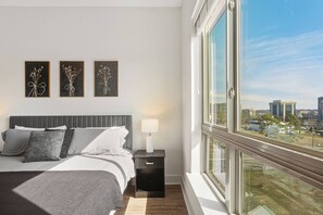 A modern bedroom with a large bed, a nightstand, and a window with a city view. - Blue Haven | Pool, City Views, Free Parking (Charlotte)