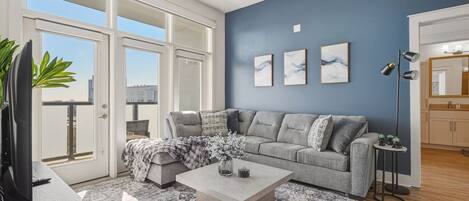 A modern living room with a grey sectional sofa, a coffee table, and a patterned rug. There are three framed artworks on the wall, a floor lamp, and a small table with plants.