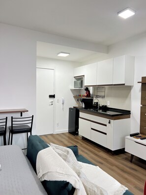 A compact kitchen with white cabinets, a microwave, and a sink. There is a dining area with a table and chairs, and a sofa with a white blanket. - Furnished studio apartment near the US Consulate, Interlagos, Morumbi Shopping Mall, and Brooklin metro station. (São Paulo)