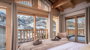 Une chambre avec un grand lit, une chaise et une vue sur des montagnes enneigées, visible à travers de grandes fenêtres.