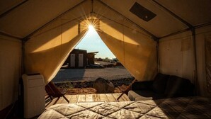 Tienda de campaña con cama, dos sillas y vista a un edificio cercano.