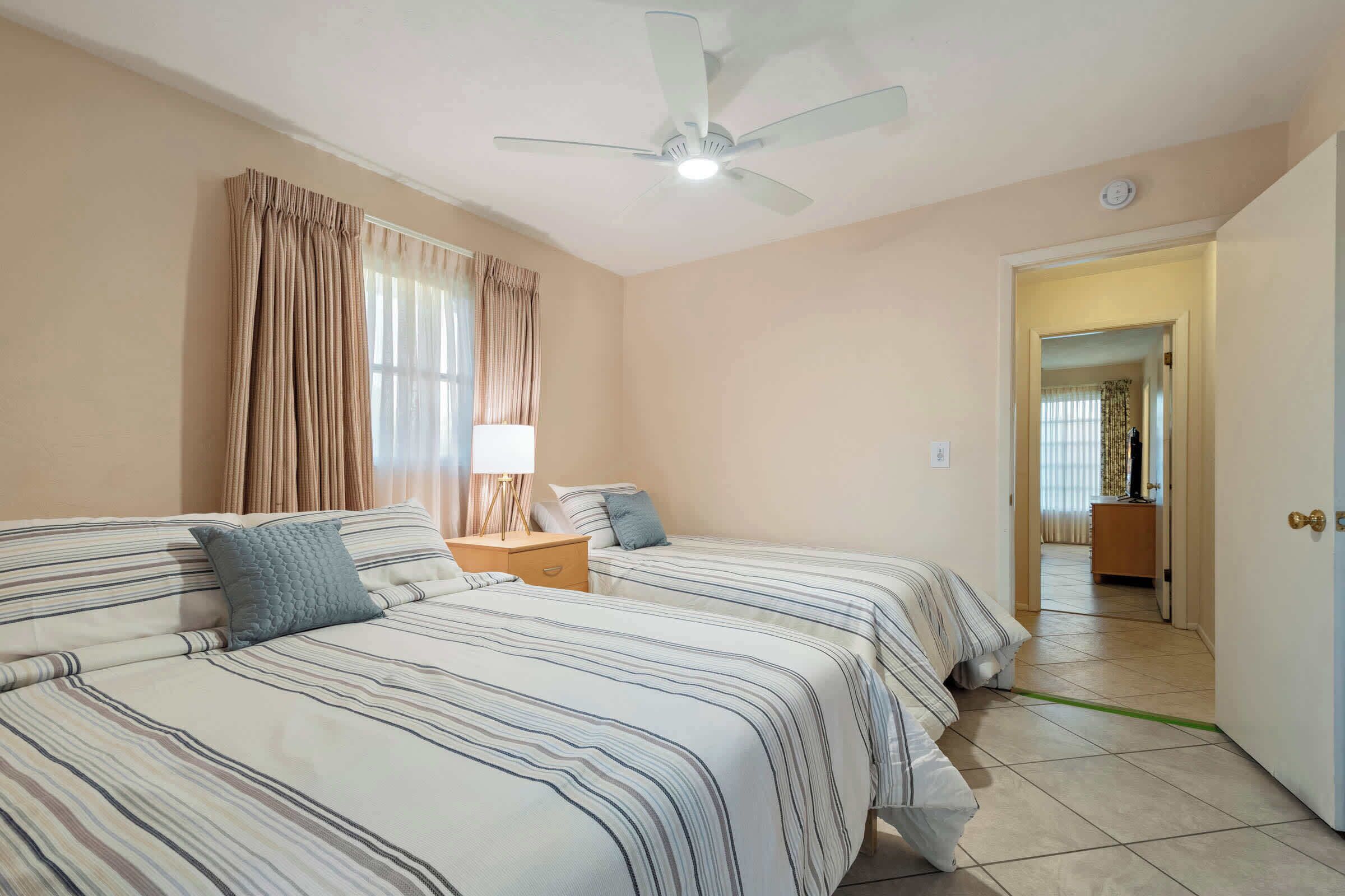 A hotel room with two beds, a ceiling fan, a nightstand, and a view into the hallway.