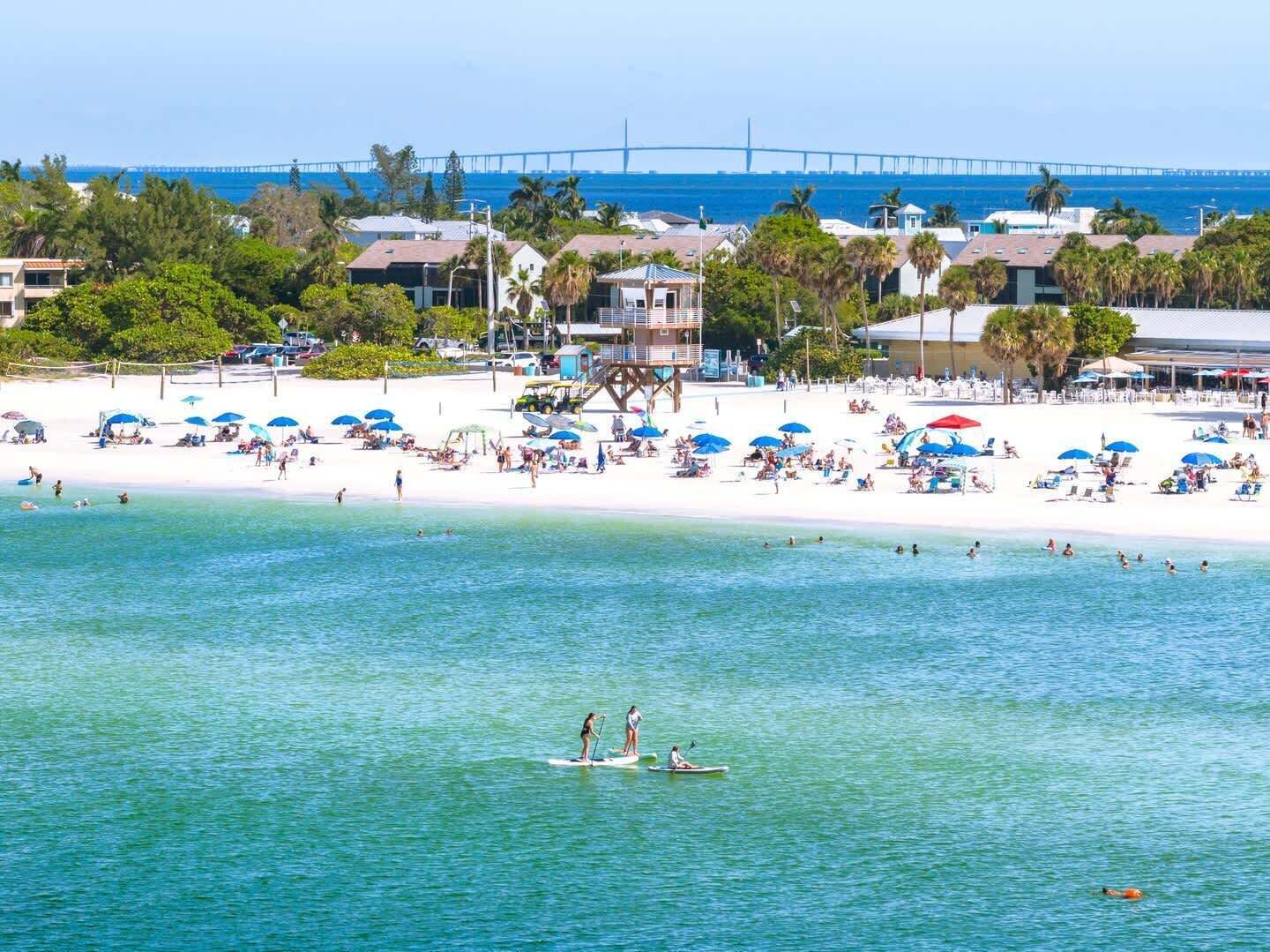 A beach with people sunbathing and swimming, a lifeguard tower, and a bridge in the background.
