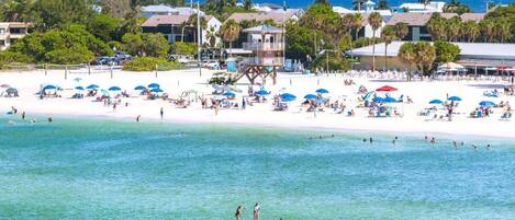 A beach with people sunbathing and swimming, a lifeguard tower, and a bridge in the background.