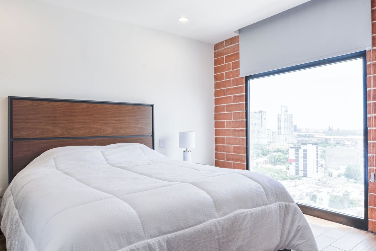A bedroom with a large bed, a wooden headboard, a white lamp, and a view of a cityscape through a large window.