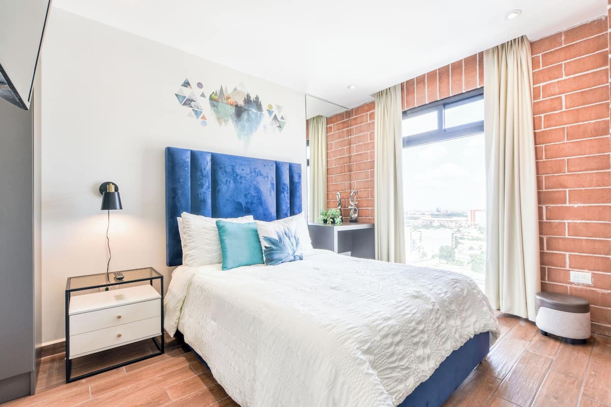 A modern bedroom with a large bed, a nightstand, a desk, and a view of the city through a large window.