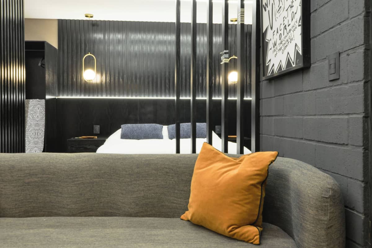 A modern hotel room with a grey sofa, a mustard-colored pillow, a bed with a blue and white duvet, and a wall-mounted light fixture.