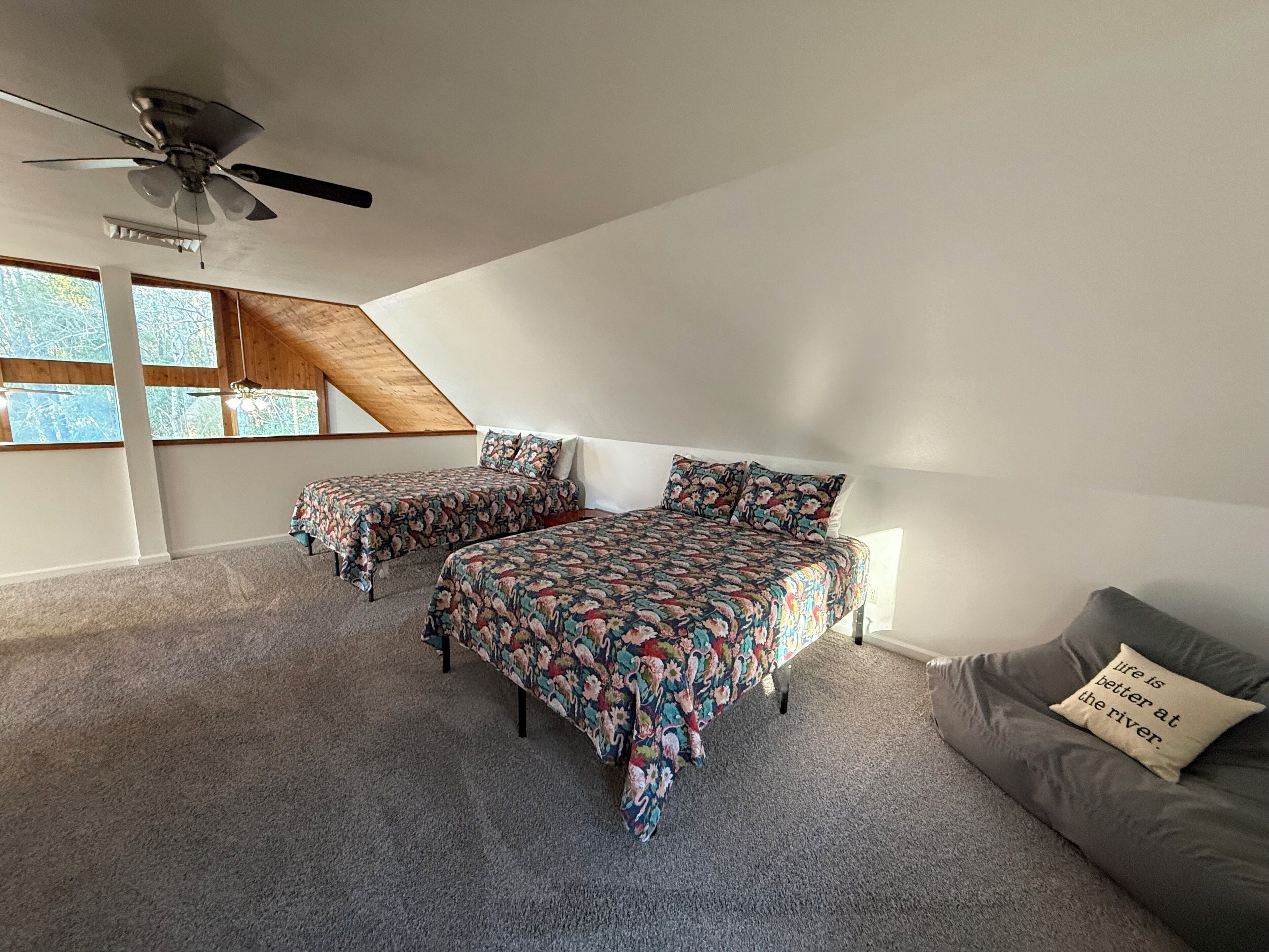 Un dormitorio con techo inclinado, ventilador de techo, dos camas con ropa de cama floral, un sofá gris con un cojín y una ventana con vistas a árboles.