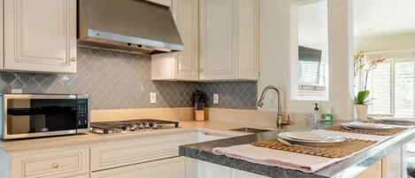 Una cocina con armarios blancos, microondas de acero inoxidable, estufa a gas y encimera de granito.