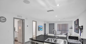 Una cocina moderna con encimera negra y pileta de acero inoxidable, un comedor con mesa y sillas blancas, y una sala de estar con sofá y televisor.
