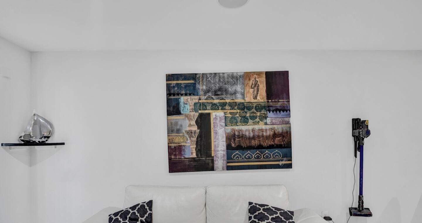 A room with a white wall, a painting, a white sofa with patterned cushions, and a floor lamp.