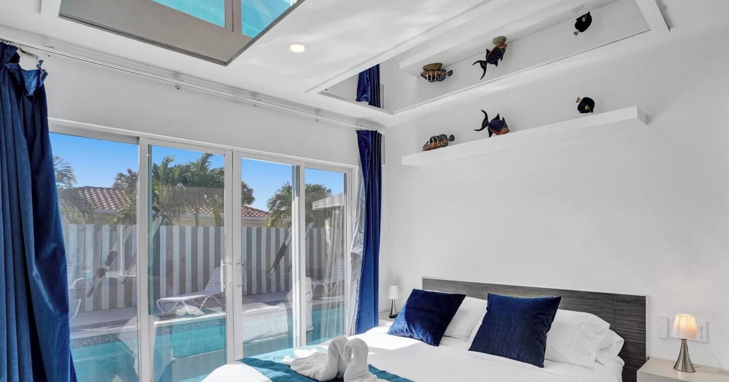 Une chambre d’hôtel comprenant un lit, une table de chevet, une lampe, une fenêtre avec des rideaux et une vue sur une piscine et un espace extérieur.