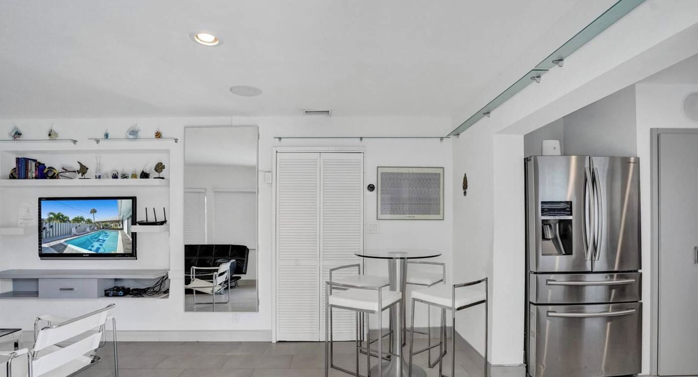 A modern kitchen with a stainless steel refrigerator, a dining area with a round table and chairs, and a TV mounted on the wall displaying a pool scene.