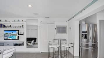 A modern kitchen with a stainless steel refrigerator, a dining area with a round table and chairs, and a TV mounted on the wall displaying a pool scene.