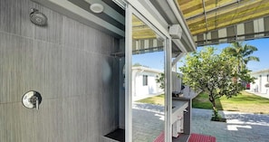 A modern bathroom with a shower, a towel rack, and a glass door leading to an outdoor area with a tree and a pathway. - Private house w/pool & patio (Deerfield Beach)
