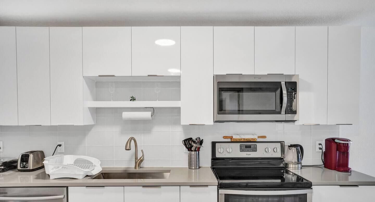 Una cocina moderna con gabinetes blancos, un lavamanos de acero inoxidable, un microondas, una estufa a gas y una cafetera roja.