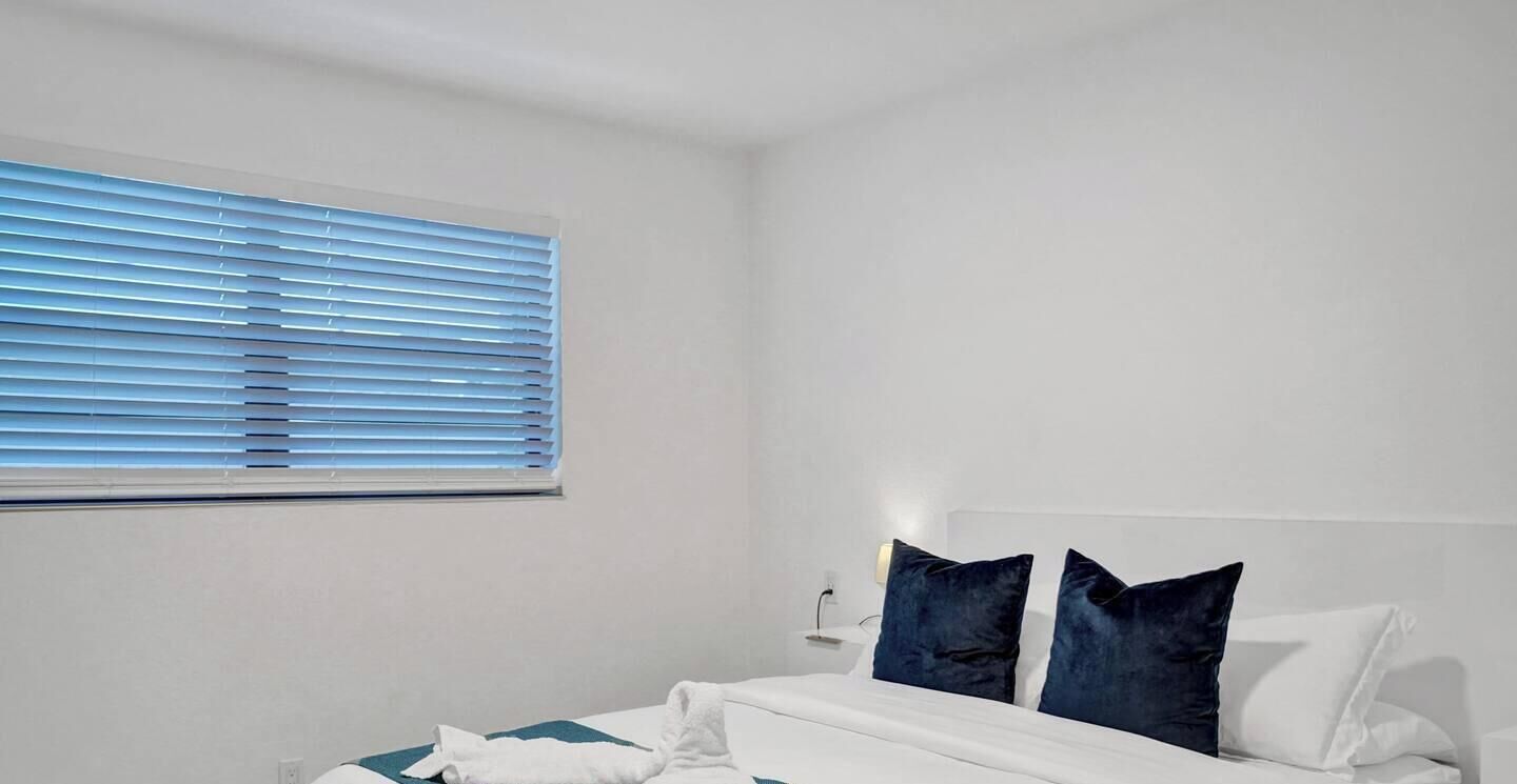 Una habitación de hotel con una cama, un ventanal con persianas y una mesita de noche con lámpara.