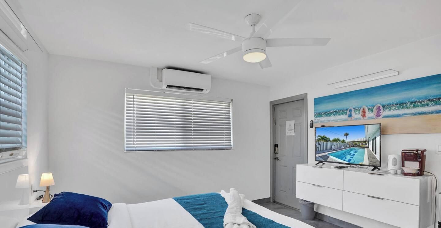 A hotel room with a bed, a desk, a ceiling fan, an air conditioning unit, a window with blinds, a door, a painting, and a television.