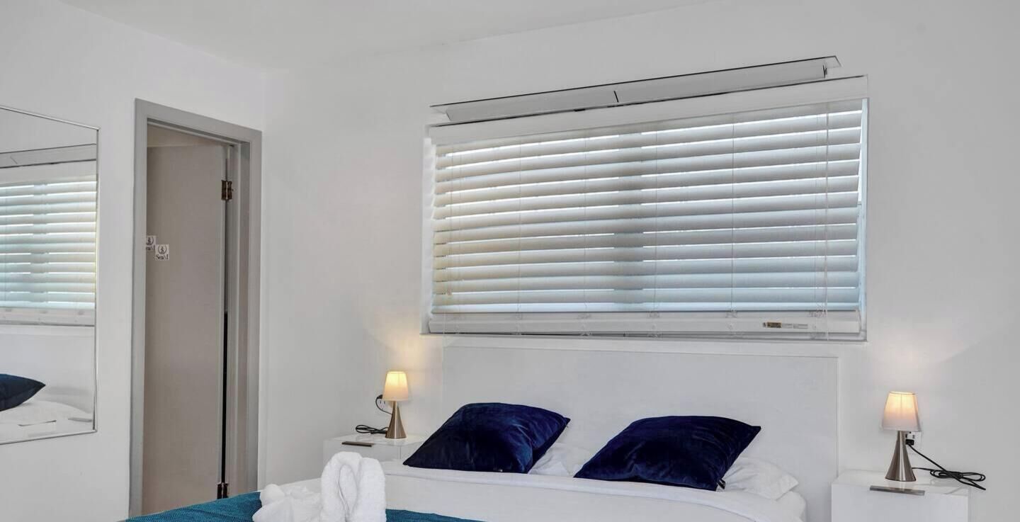 A hotel room with a bed, two bedside tables with lamps, a window with blinds, and a door.