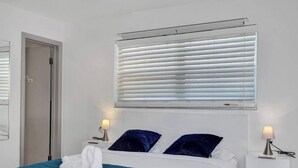 Une chambre d’hôtel avec un lit, deux tables de chevet avec des lampes, une fenêtre avec des stores et une porte.