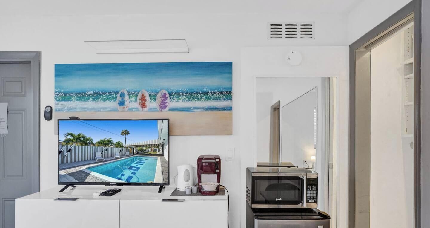 A room with a flat-screen TV displaying a poolside view, a coffee maker, a microwave, and a painting of surfboards on the wall.