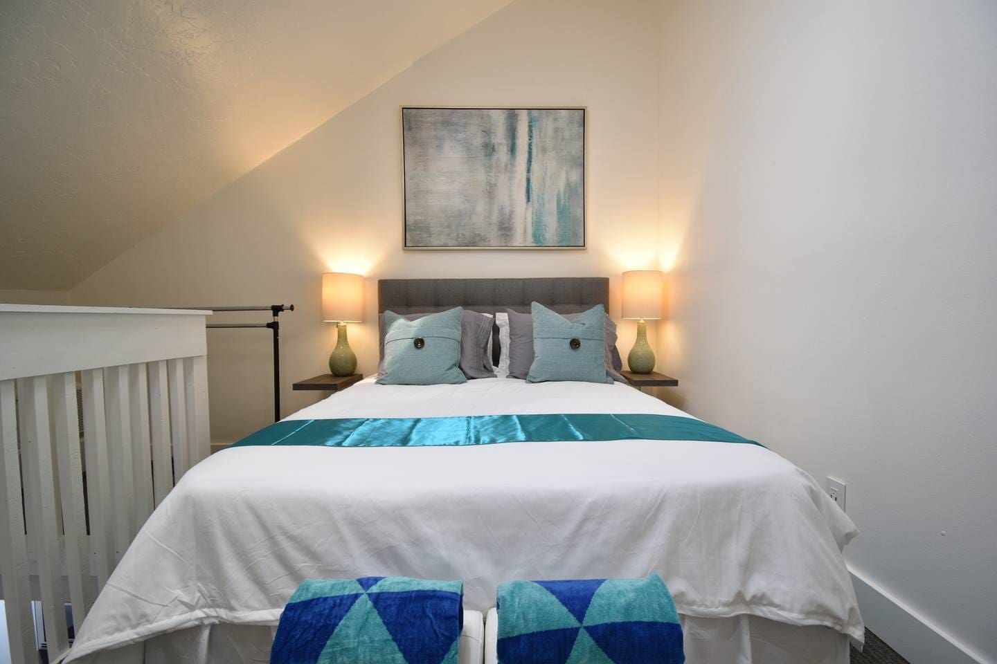 A bedroom with a bed, two bedside lamps, a wall-mounted painting, and a window with blinds.