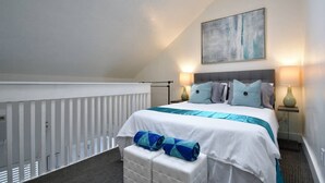 Ein Schlafzimmer mit Bett, Kissen, einem Nachttisch, einer Lampe, einem Bild an der Wand und einem weißen Geländer.