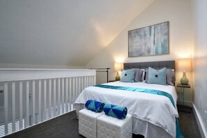 Een slaapkamer met een bed, kussens, een nachtkastje, een lamp, een schilderij aan de muur en een witte balustrade.