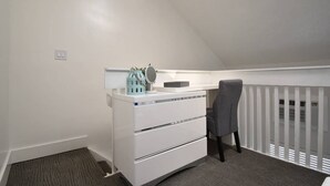 Une petite chambre de style « loft », aux murs blancs, comprenant un bureau et une chaise, une commode et une fenêtre avec des stores.