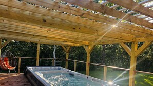 Un bain à remous installé sous une pergola en bois, avec une terrasse en bois et un fauteuil suspendu.