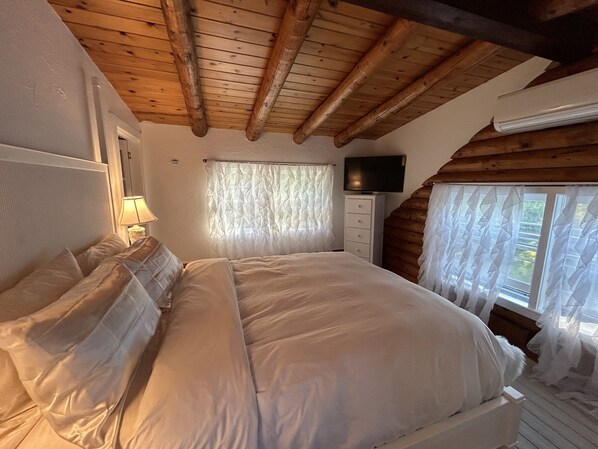 Un dormitorio con techo de madera, cama con ropa de cama blanca, televisor en la pared y ventana con cortinas de gasa.