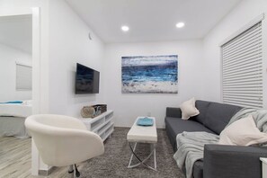 Ein modernes Wohnzimmer mit einem grauen Sofa, einem weißen Sessel, einem Couchtisch und einem Gemälde mit einer Strandansicht.