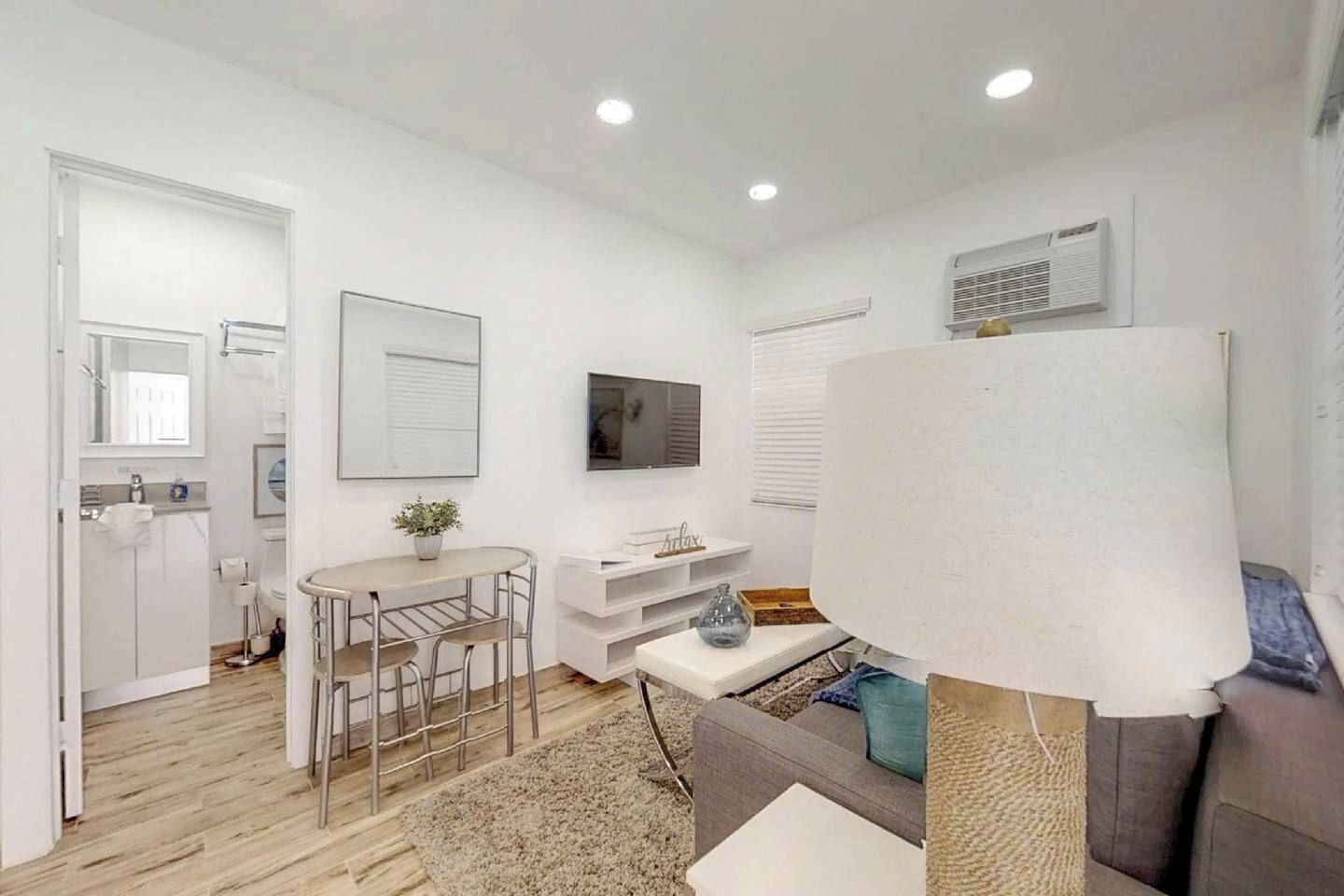 A modern, well-lit room with a dining area, a sofa, a TV, and a window with blinds.