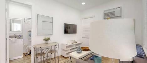 A modern, well-lit room with a dining area, a sofa, a TV, and a window with blinds.