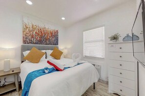 A hotel room with a bed, bedside lamps, a painting, a window with blinds, a dresser, and a television. - Modern studio w/queen bed & beach views (Hollywood)
