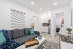 A modern apartment with a grey sofa, a small wooden tray, a lamp, and a kitchen area visible through an open door. - Modern studio w/queen bed & beach views (Hollywood)