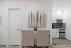 Ein minimalistisch eingerichteter Raum mit einer weißen Tür, einer Sitzecke mit zwei Sesseln, einem an der Wand befestigten abstrakten Gemälde und einer Küchenzeile mit Mikrowelle und Kühlschrank.