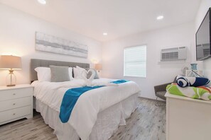 A hotel room with a bed, bedside table, lamp, air conditioning unit, window, and a chair. - Modern apartment w/king bed & beach views (Hollywood)