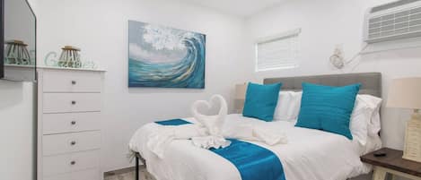 A hotel room with a bed, a dresser, a painting, a window, and an air conditioning unit.